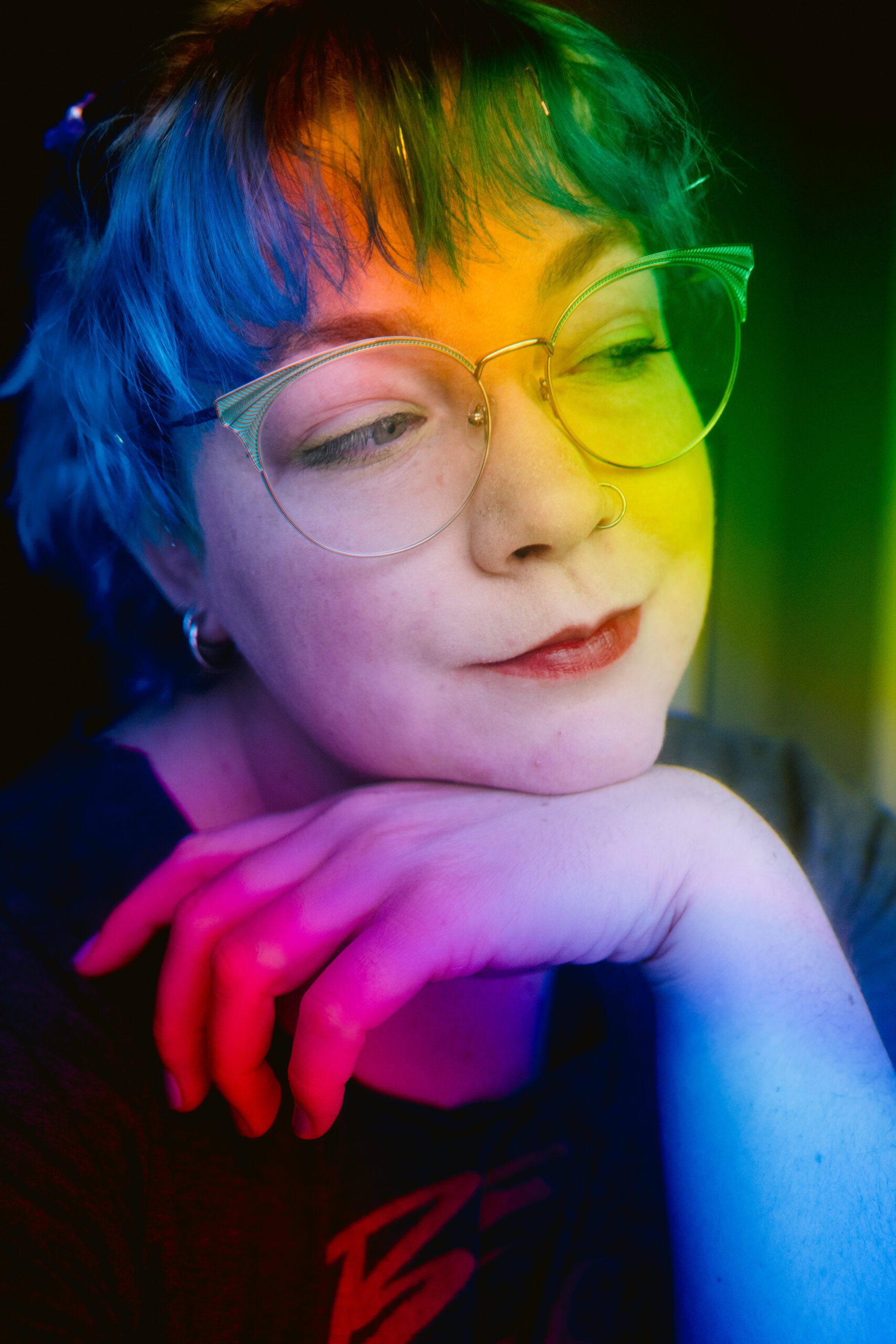 Portrait of a nonbinary subject with blue hair, glasses, and red lipstick. Their head is resting on their hand and they are looking away off camera. There is a rainbow effect over the entire image.