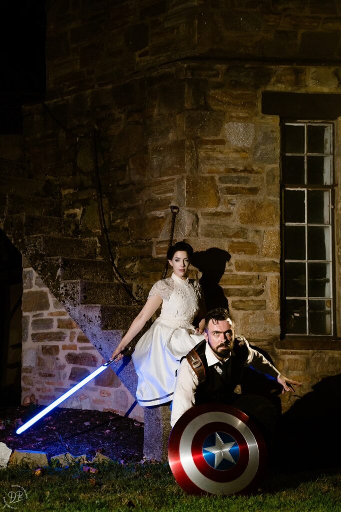 A newlywed couple posing in front of a windmill, one with a lightsaber and one with a Captain America shield 