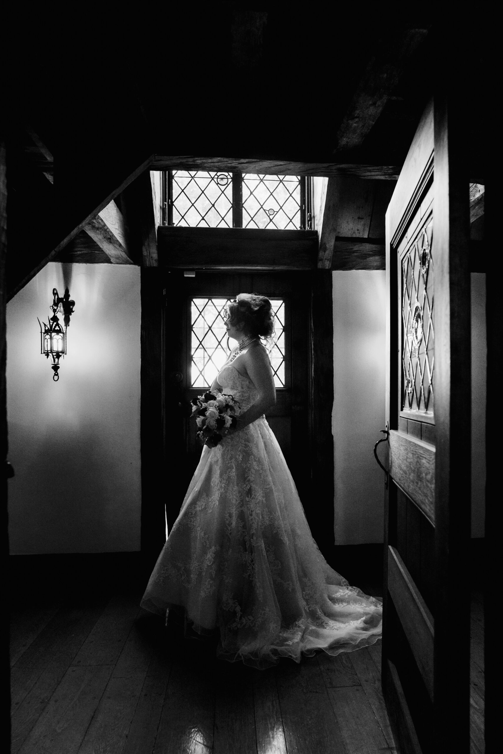 black and white photo of a woman in a wedding dress holding a boquet of flowers standing in a hallway 