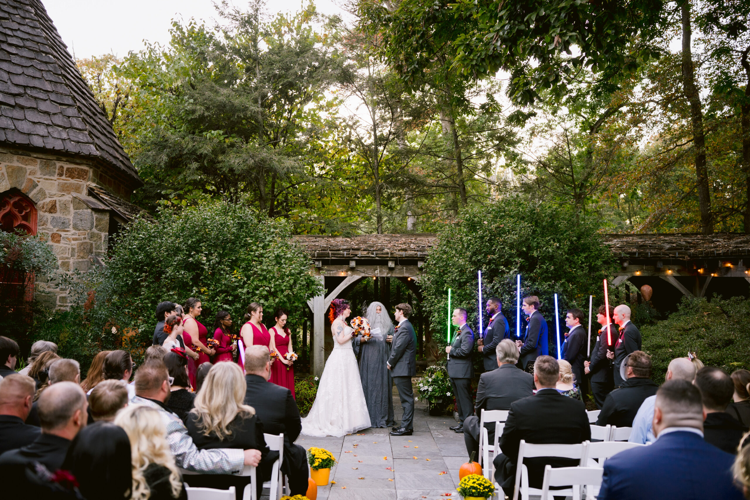 wedding ceremony with a woman in a wedding dress and her wedding party and a man with a tuxedo with his wedding party who are all holding light sabes while guests sit in chairs watching the ceremony