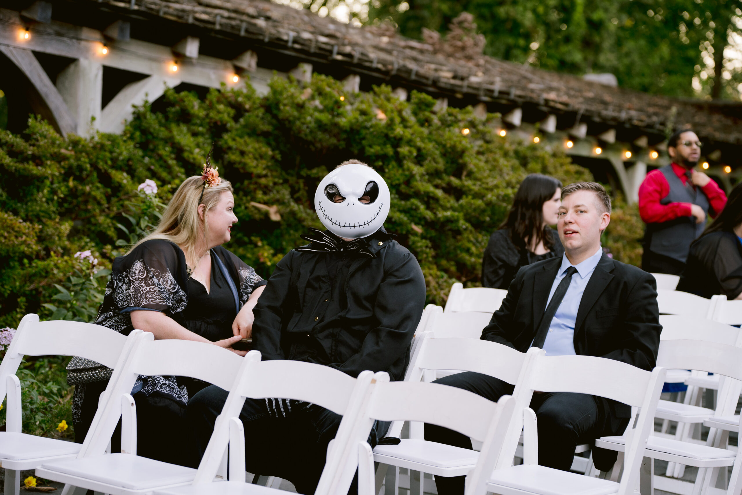 wedding guests sitting in white chairs with a man dressed in a halloween costume 