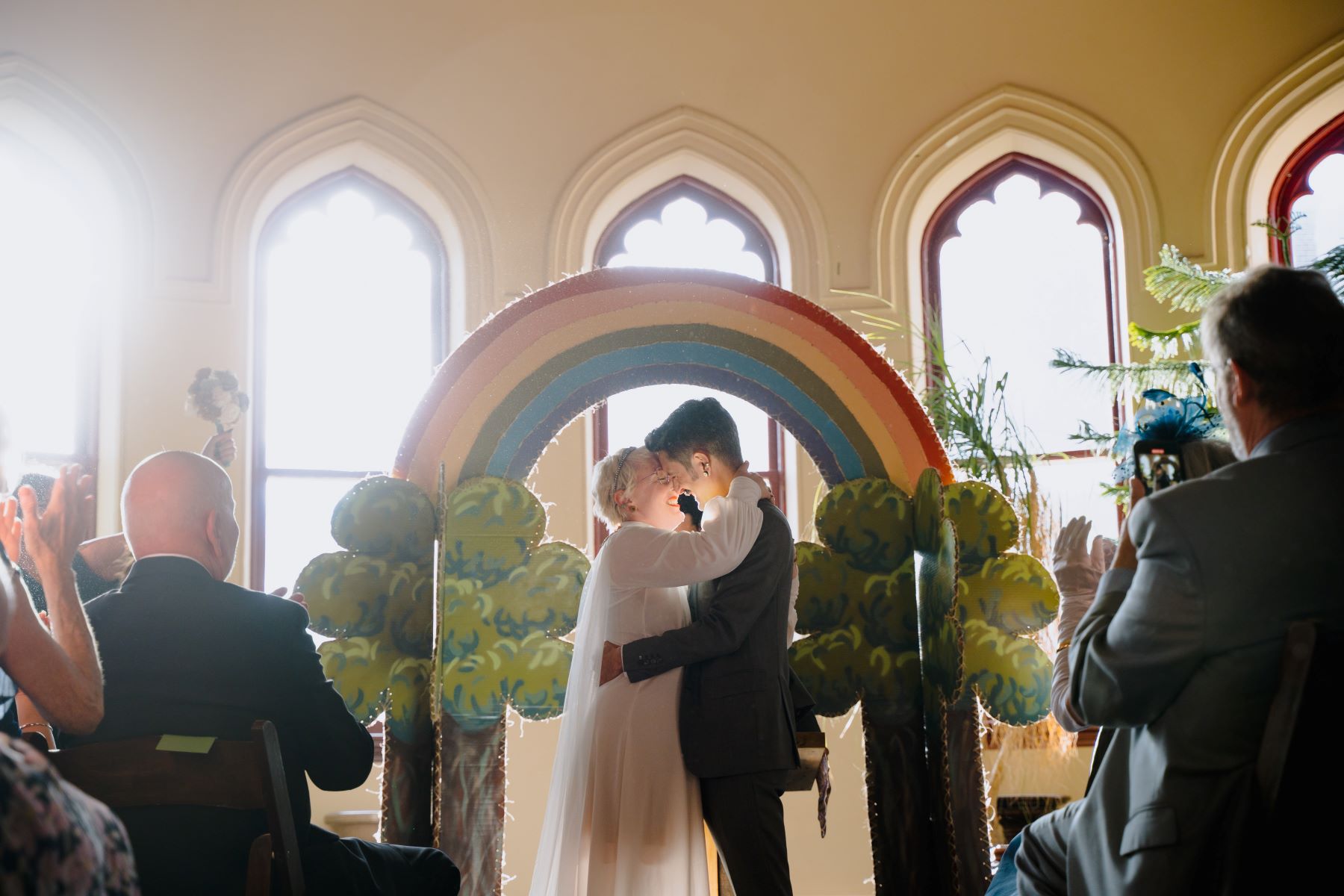 A couple kissing in front of a rainbow arch at their wedding surrounded by their guests who are cheering for them one is wearing a white dress and the other is wearing a black suit