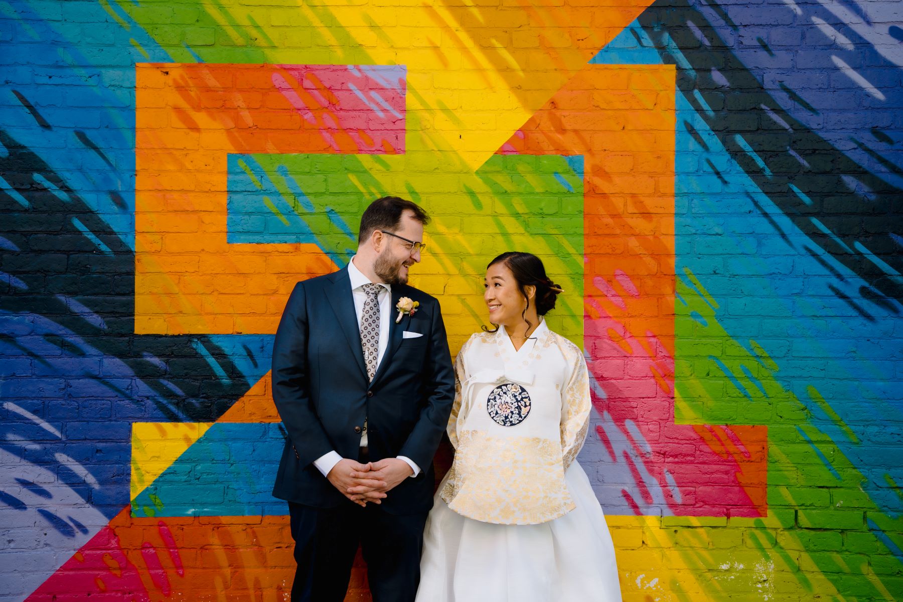 A woman in a white wedding kimono smiling at her partner who is also looking at her and smiling adn waring a black suit, a white button up, a tie, glasses, and a boutonniere