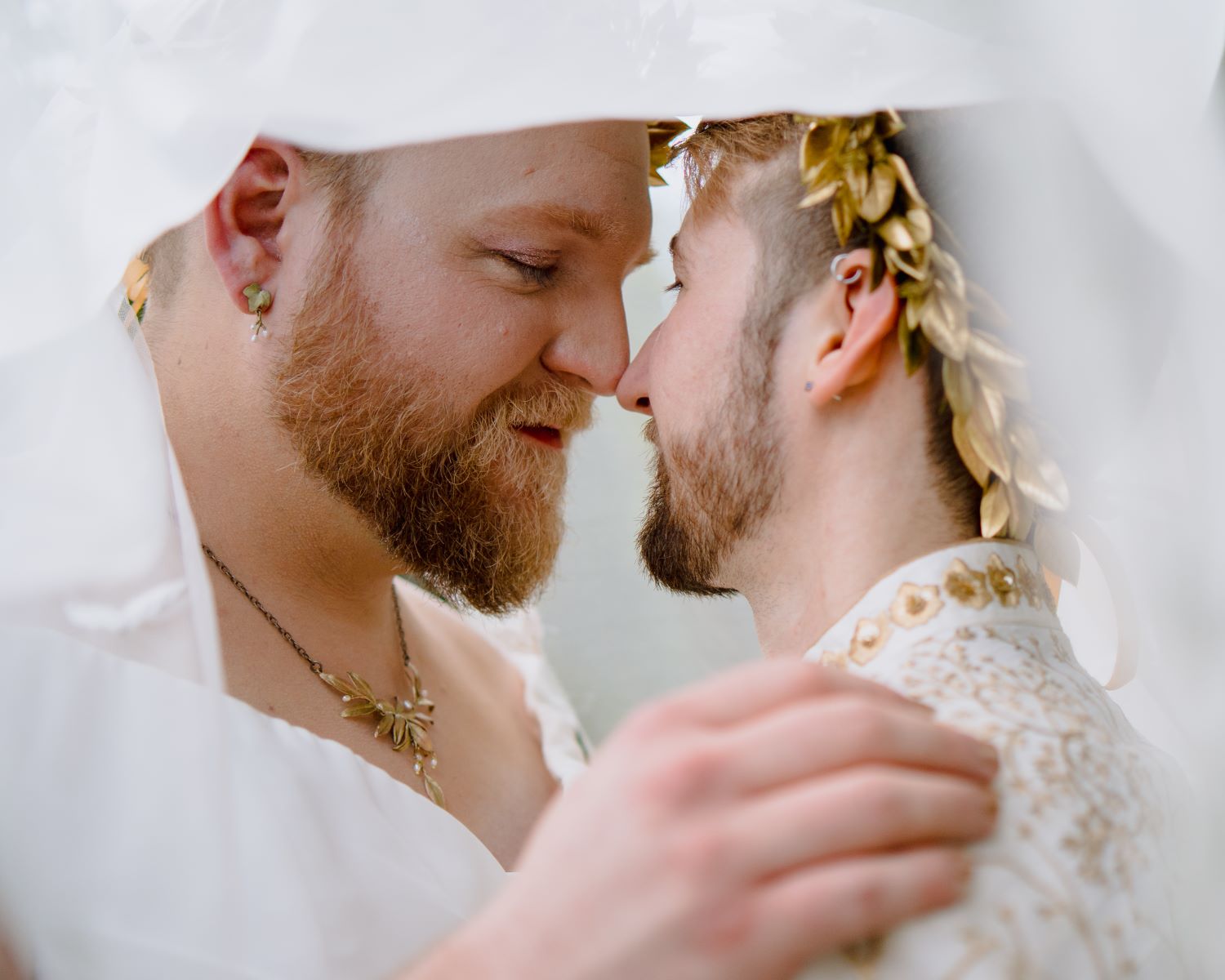 Couple with their noses pressed together and both are smiling both have earrings and both are wearing gold leaf crowns and white attire 