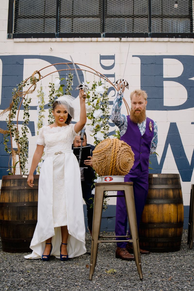 A man and woman with a sword and in front of them is a stool with a ball of twine to cut the woman is wearing a white wedding dress a white birdcage veil and the man is wearing a purple vest with purple pants and a patterned blue button up underneath behind them are barrels and a wedding officiant 