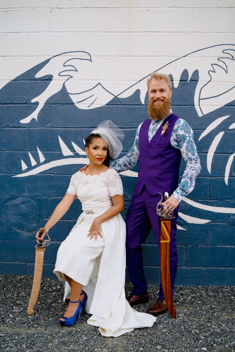 A woman with a white dress sitting down wearing a birdcage white veil and blue heels and posing with a sword and her partner is touching her shoulder and wearing a purple vest and purple pants and a patterned blue and white button the man is smiling and also has a sword and the woman is softly smiling 