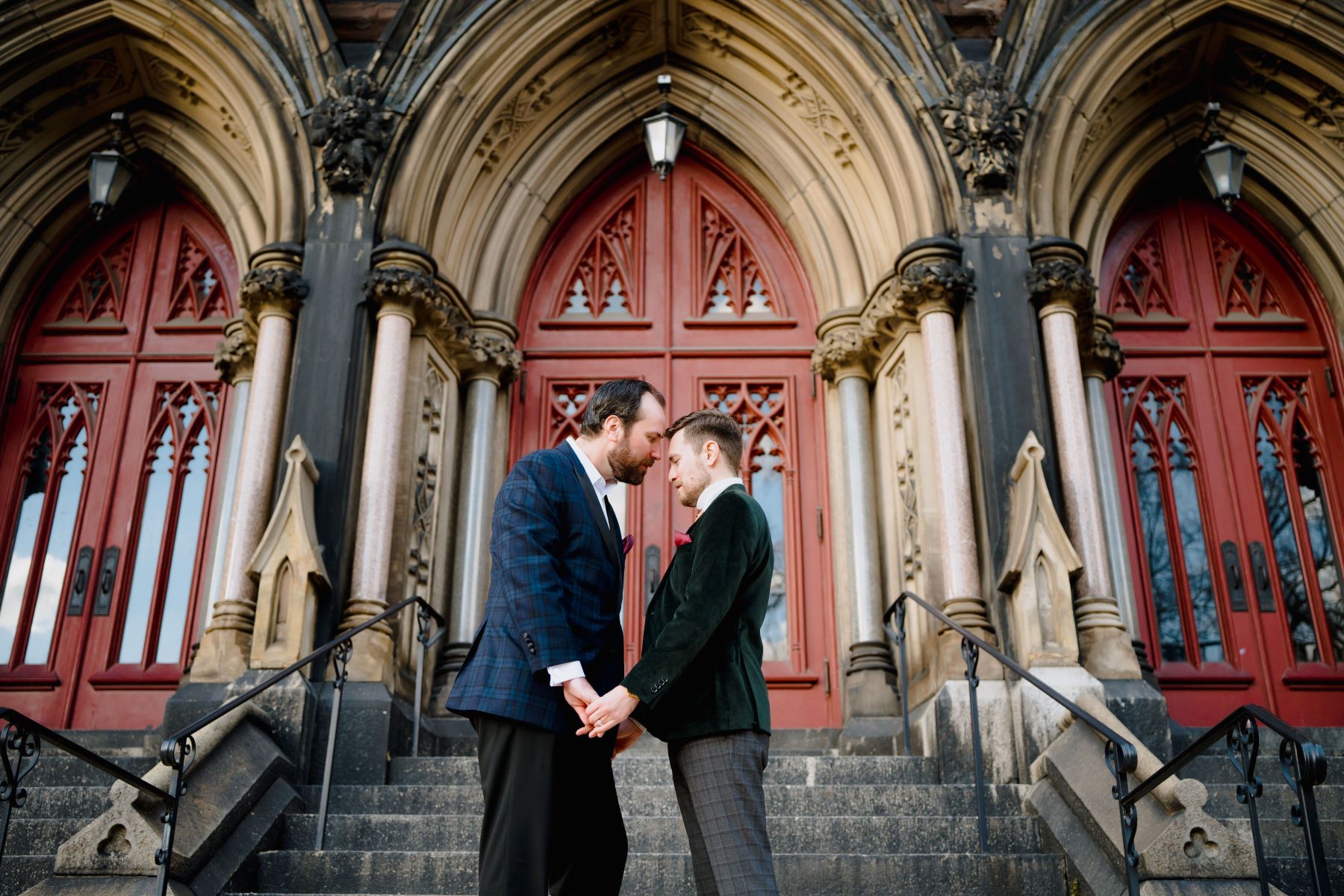 Couple standing in front of a building with gothic architecture at the bottom of a set of stairs theya re both leaning towards each other and holding hands and they both have on dress jackets and slacks 