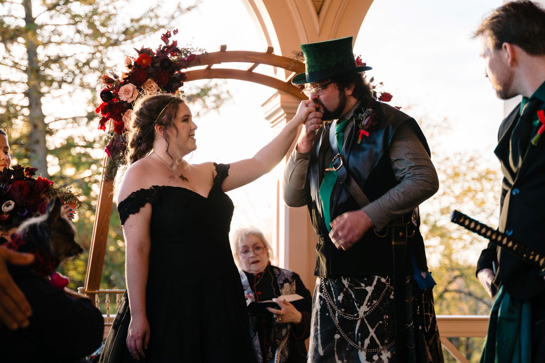 Man with a top hat and a kilt kissing the hand of the woman in front of him who is wearing a black dress an behind them is a wedding officiant and an arch