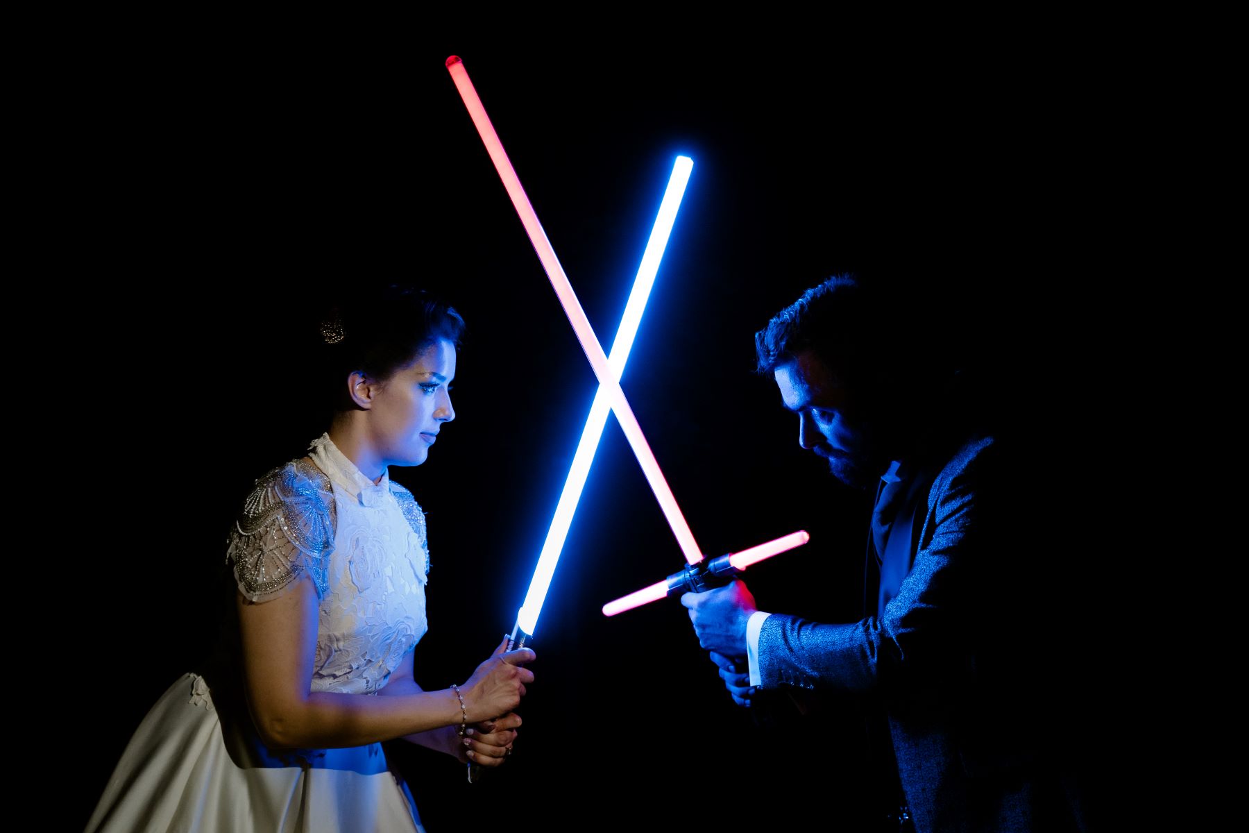 A couple playing with blue and red light sabers the woman is wearing a white dress and the man is wearing a suit 