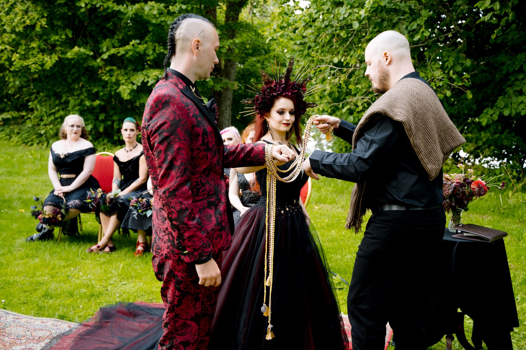 A couple and their officiant one is wearing a red and black suit and the other is wearing a crown and a red and black dress the officiant is wrapping cords around their hands and behind them are their wedding guests 