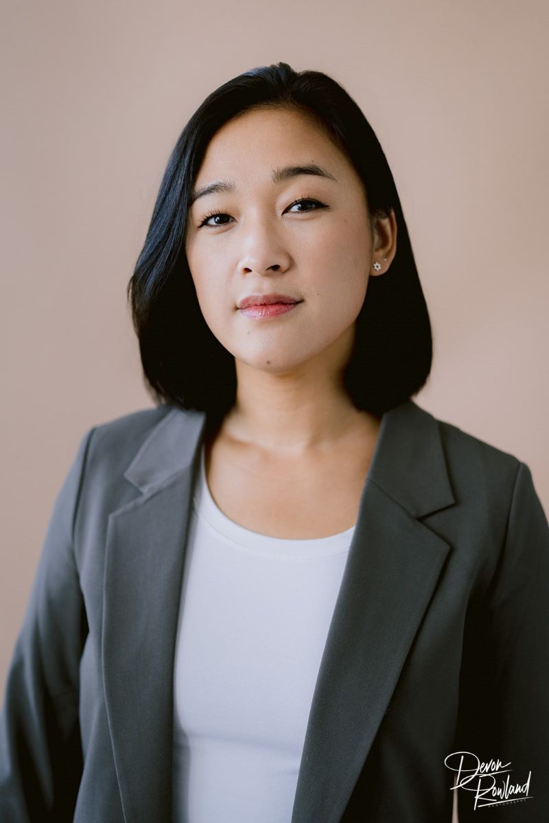 A Korean woman wearing a gray blazer and white t-shirt looking toward the camera with a beige background behind her 