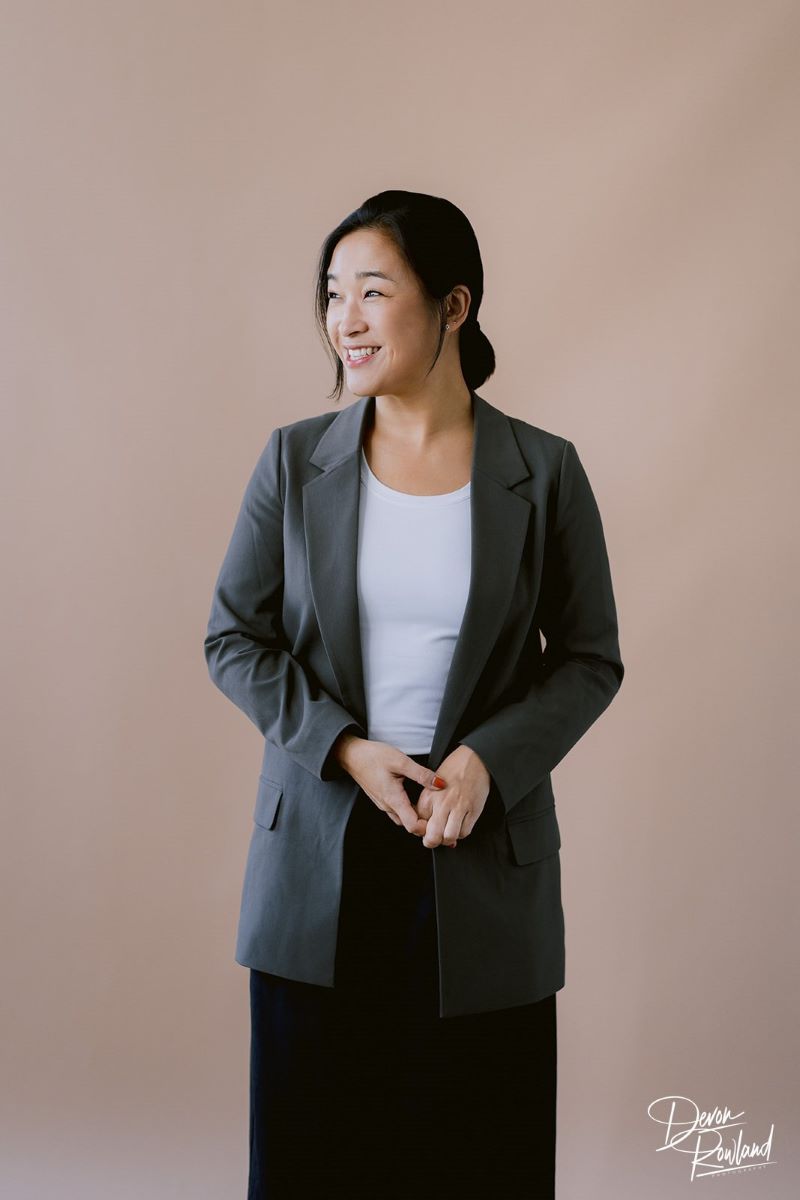 A Korean woman wearing a gray blazer, a white t-shirt, and black slacks looking to the side and and smiling with a beige background behind her 