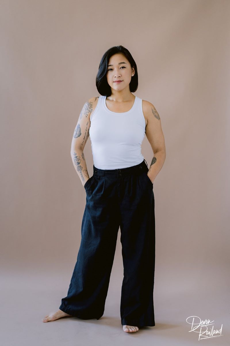 A Korean woman with tattoos in a white tank top and black slacks looking toward the camera and softly smiling with her hands in her slack pockets