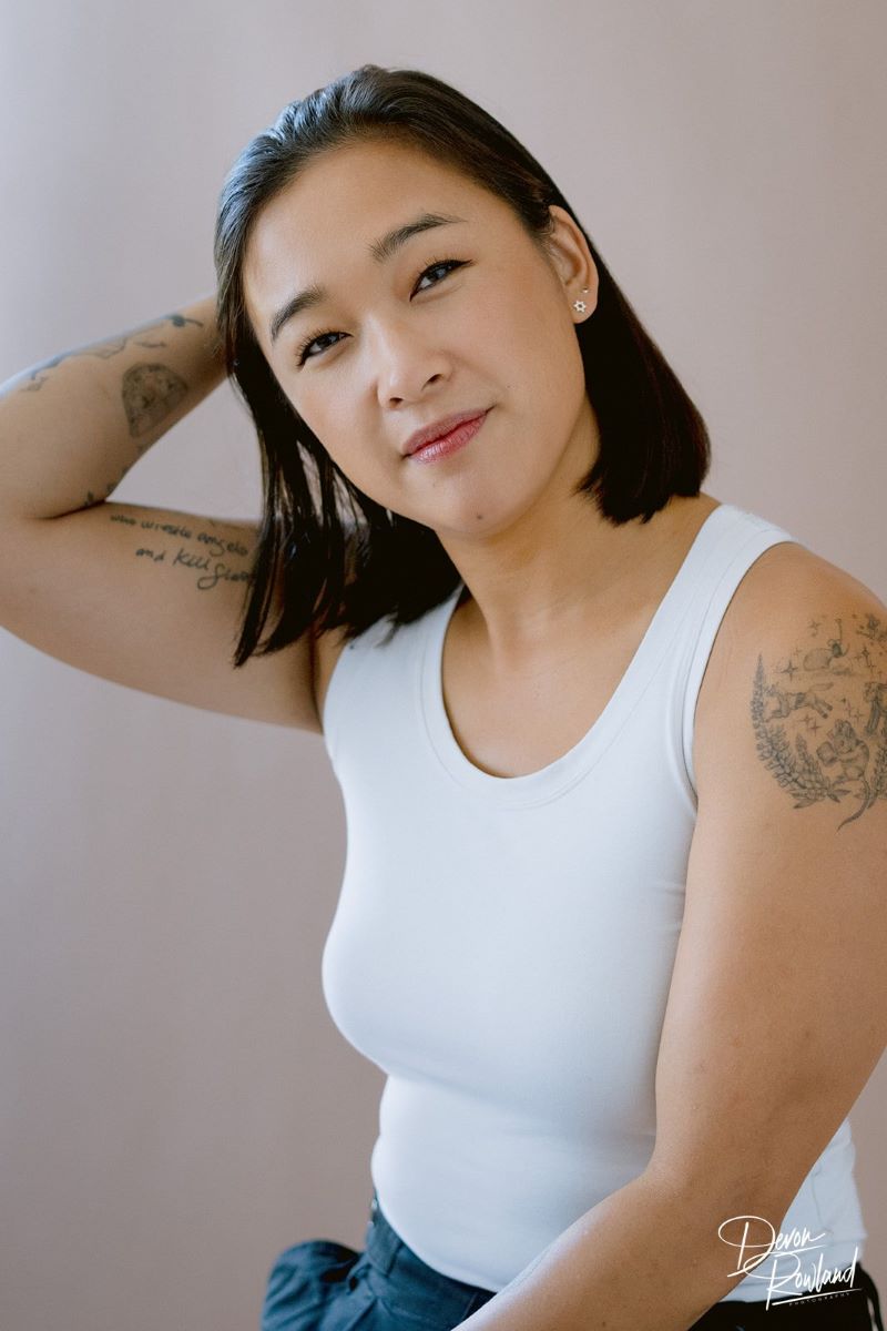 A Korean woman with tattoos wearing a white tanktop and looking toward the camera smiling softly with her head tilted and one of her hands in her hair 