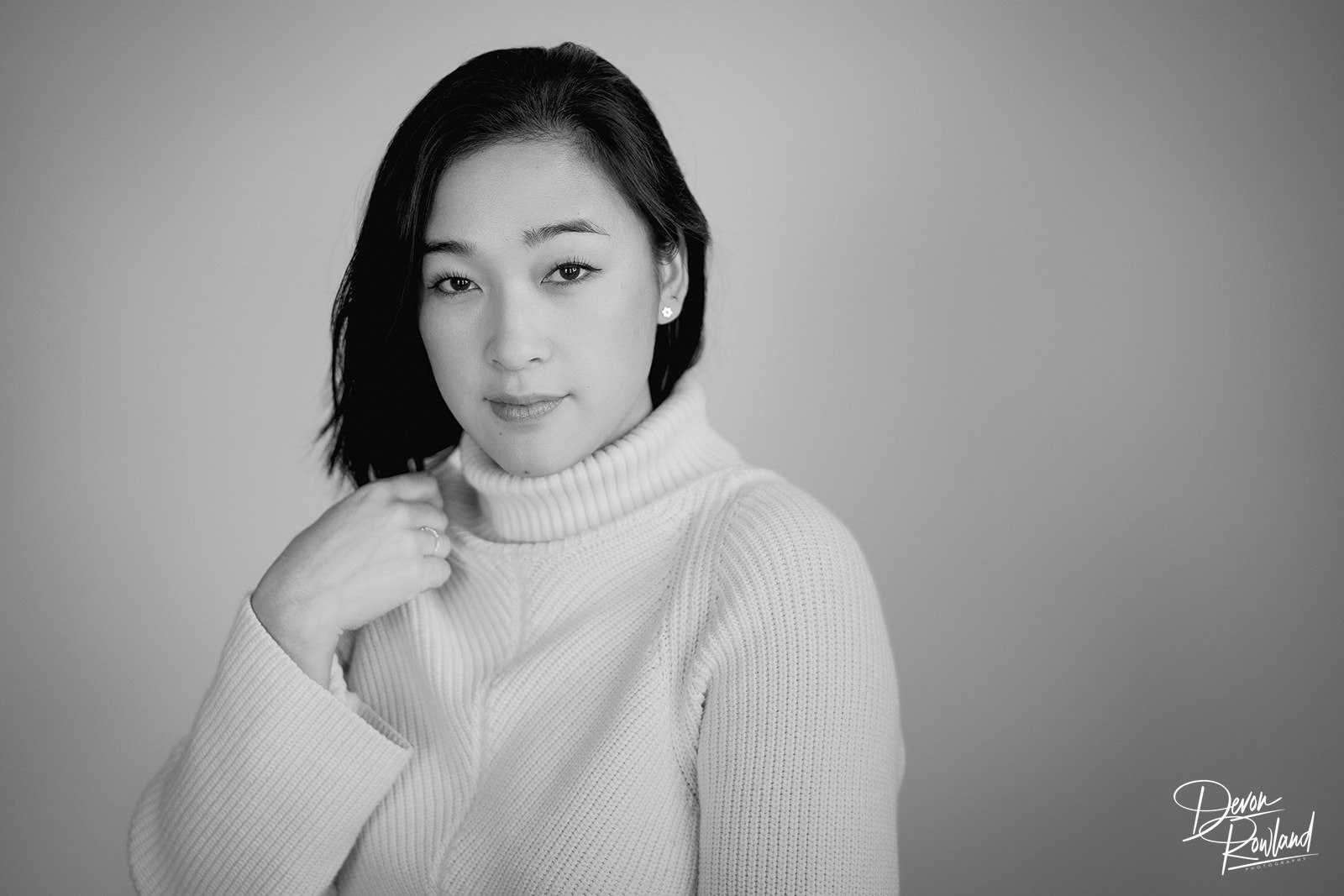 A Koorean woman in a turtleneck sweater looking toward the camera and with one hand she is lightly touching her shoulder 