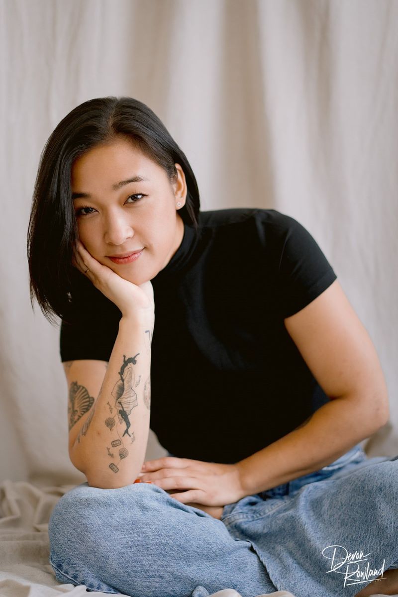 A Korean woman with tattos wearing a black t-shirt and blue jeans propping her hand underneath her chin and sitting leaning slightly to the side a cream colored plain background is behind her