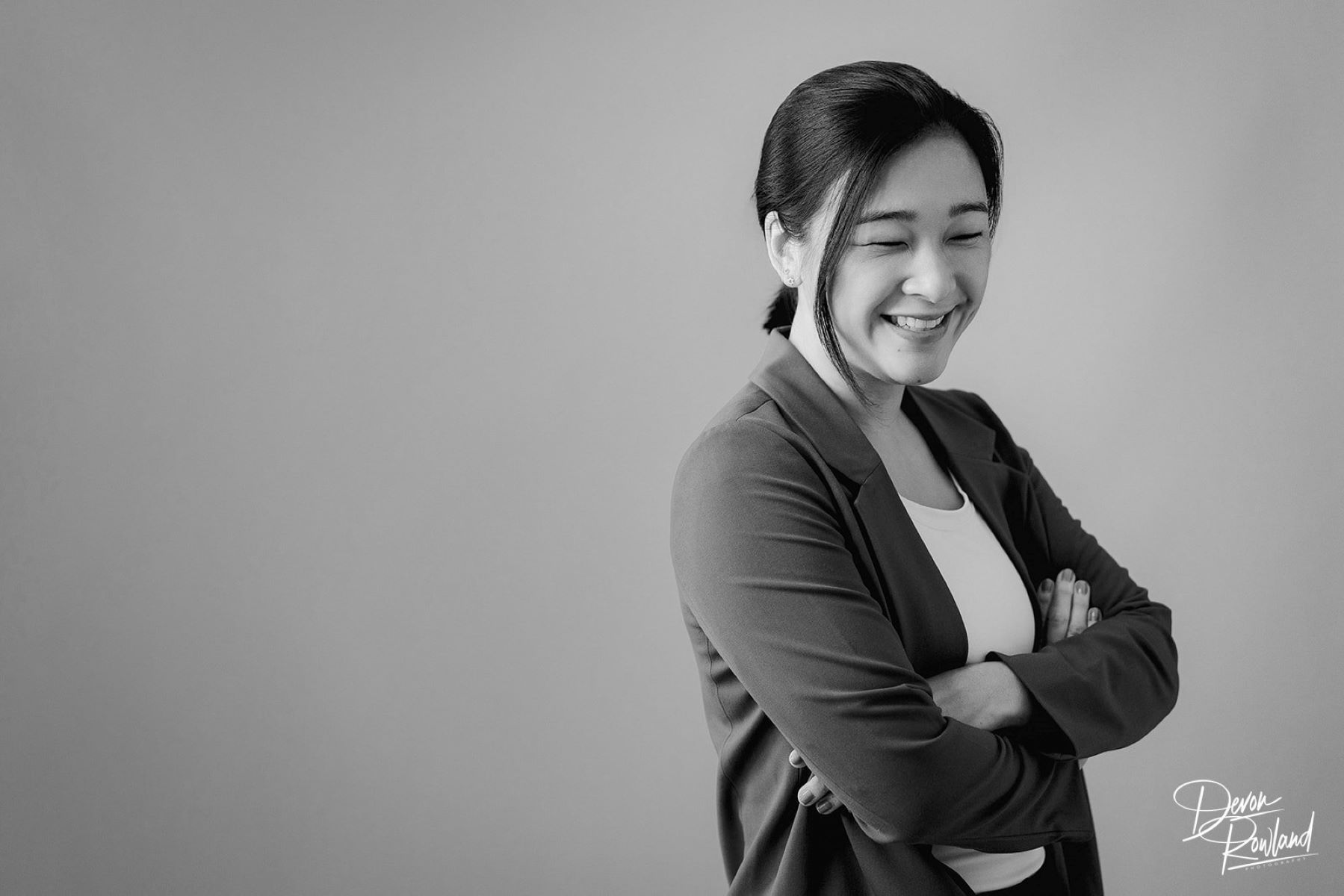 A Korean woman in a white shirt and grey blazer looking to the side smiling standing with her arms crossed 