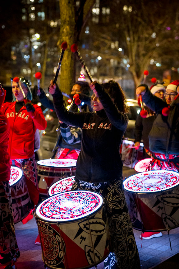 Batala DC drum troop with raised arms and drum sticks on the street at night in DC. Several members are wearing Santa hats and other holiday attire.