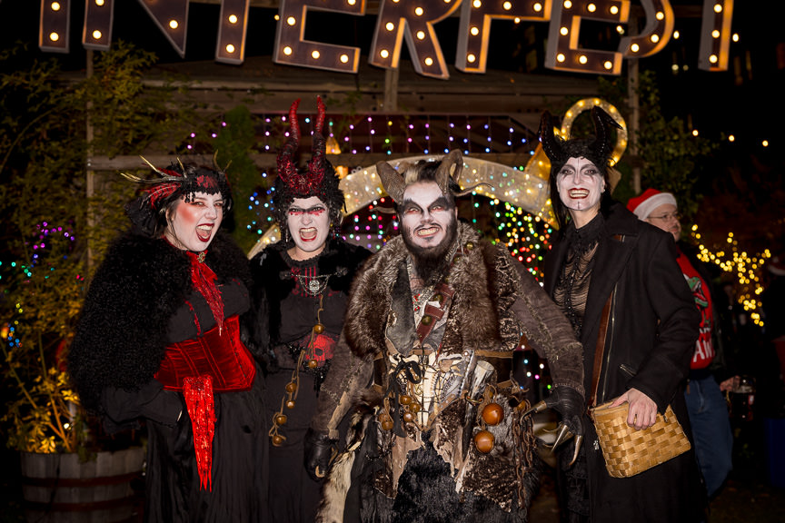 Four people wearing horns and furs for various Krampus outfits grin and grimace for the camera while standing in front of holiday lights.