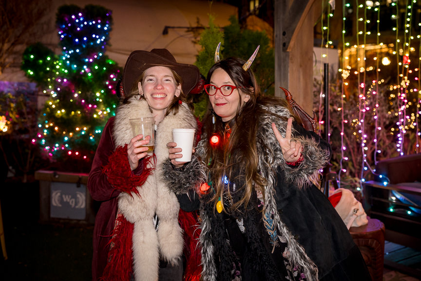 Two women wearing furs and horns hold drinks while posing for the camera.