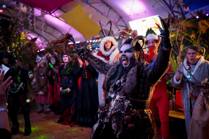 Horned Krampus stands in front of a line of various Krampuses wearing furs and making faces for the crowd while waving a flogger during the Krampus costume contest.