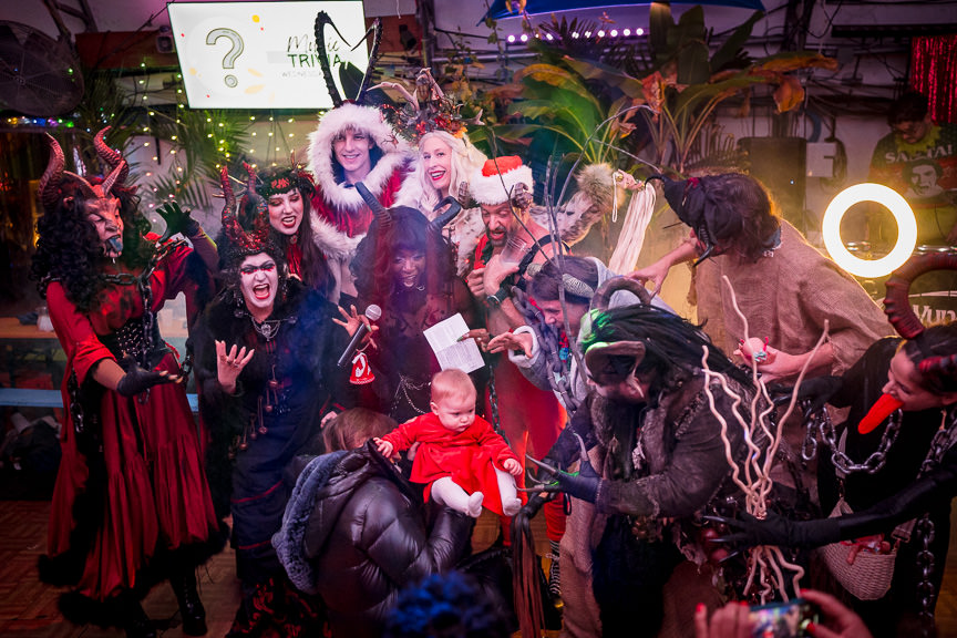 A horde of people dressed in various Krampus costumes surround a baby in a red dress for a photo opportunity.