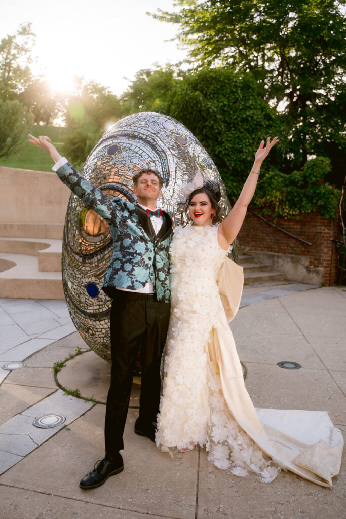 A newlywed couple with their arms up celebrating in front of an art sculpture 