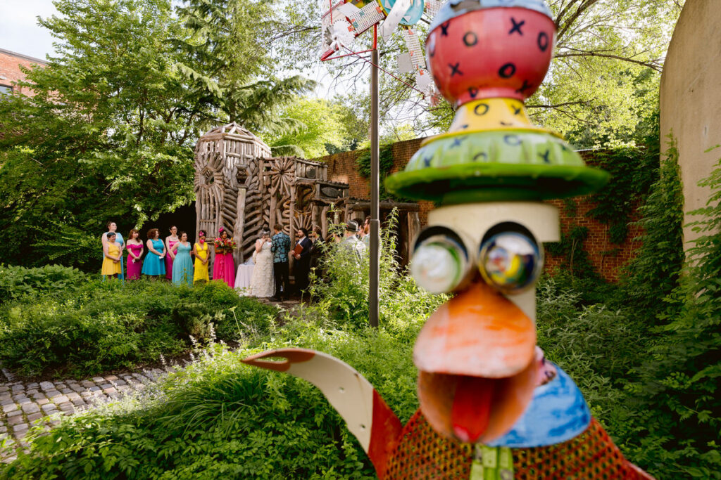 A colorful art sculpture in a garden with a wedding ceremony happening in the background 