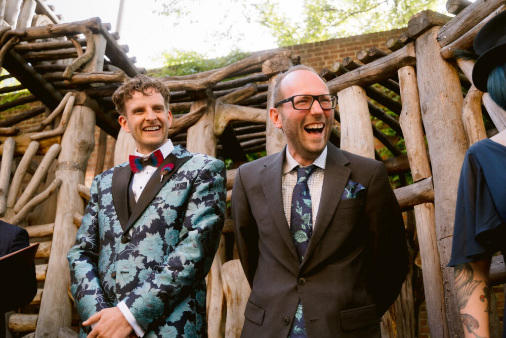 A person in a wedding suit standing up during their ceremony laughing with another member of their wedding party. 