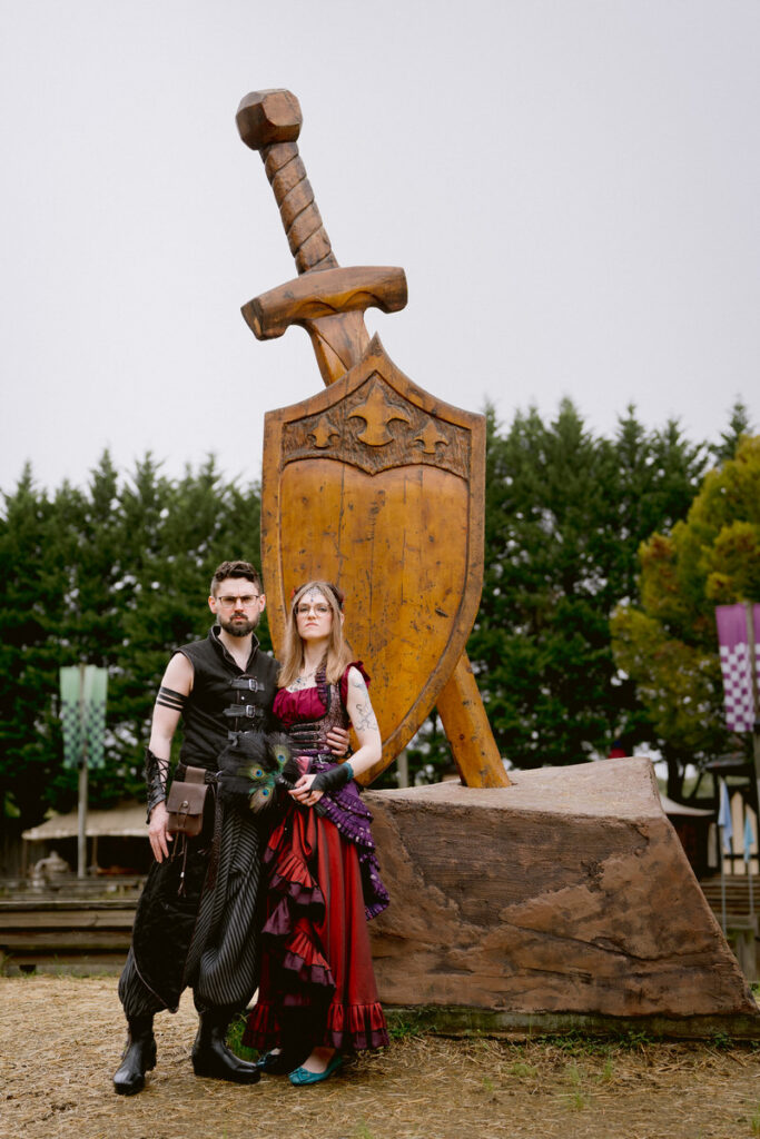 A newlywed couple posing in front of a large, wooden sword and shield at their Renaissance Festival wedding 