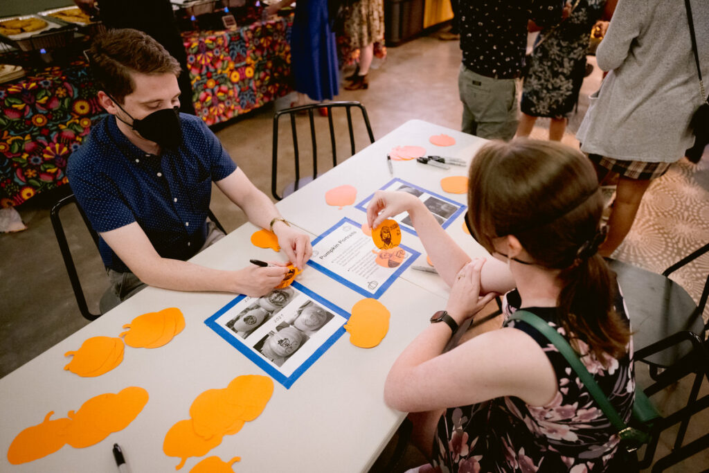 People sitting at a table drawing self-portraits on paper pumpkins 
