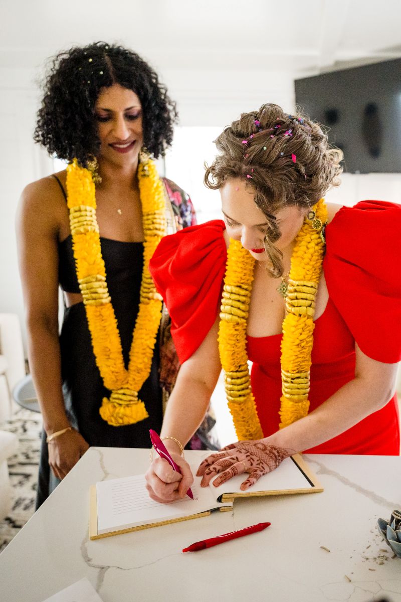 a couple inside of a boat one woman is wearing a red dress and yellow lei and is writing in a notebook the other woman is wearing a black dress and yellow lei and watching her partner 