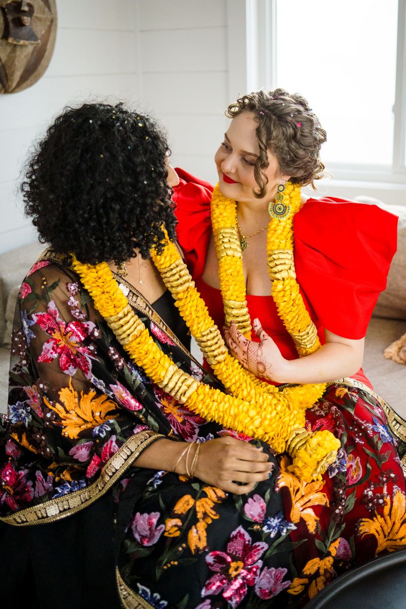 a couple sitting on a couch looking at each other one woman is wearing a red dress and yellow lei her partner is wearing a black dress and a kimono 