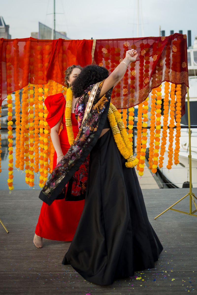 a couple dancing on a deck on the harbor one woman is wearing a black dress a kimono and a yellow lei the other woman is wearing a red dress and a yellow lei behind them is they wedding arch which is decored with yellow and orange elements 