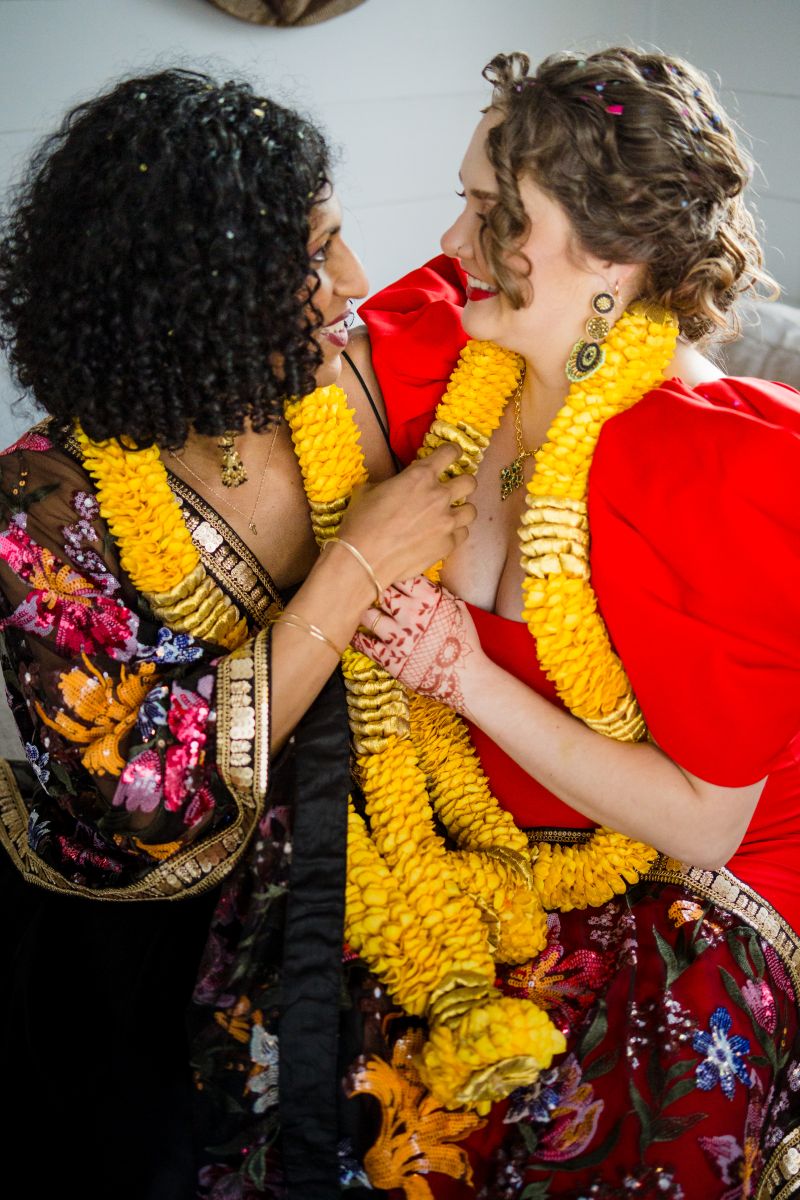 a couple sitting on a couch together smiling and hugging each other one woman is wearing a red dress and yellow lei the other woman is wearing a kimono and yellow lei 