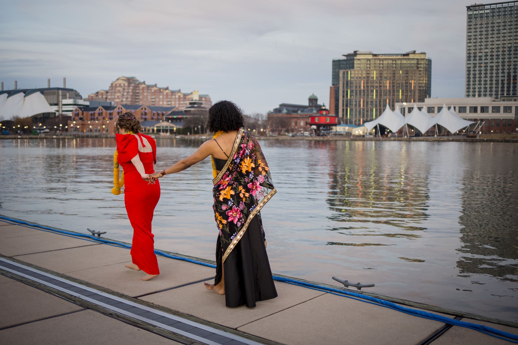 a couple walking barefoot along the harbor and holding hands the woman leading is wearing a red dress and yellow lei her partner who is following her is wearing a black dress and colorful kimono