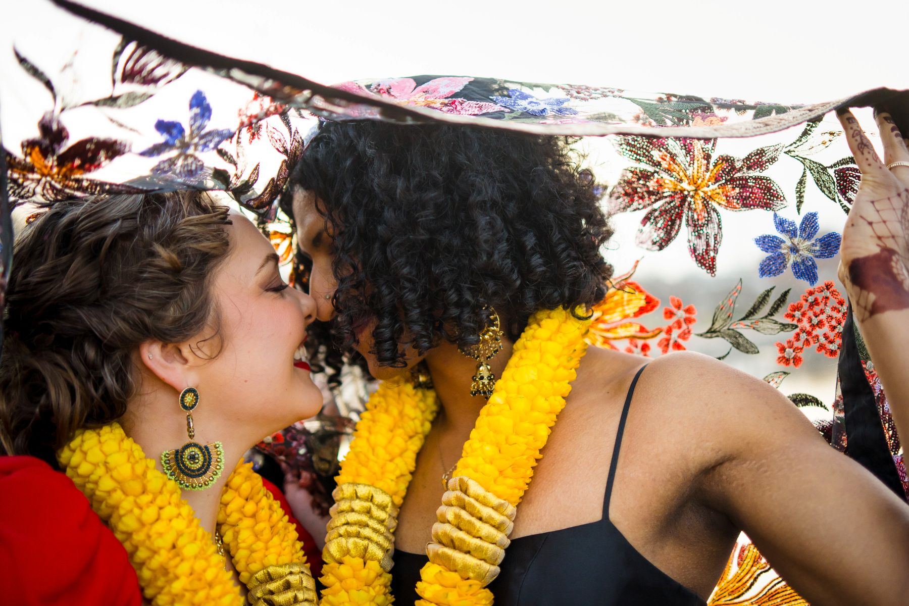 a couple looking at each other and smiling one woman is holding up a colorful floral kimono over their heads and both women are wearing yellow leis 