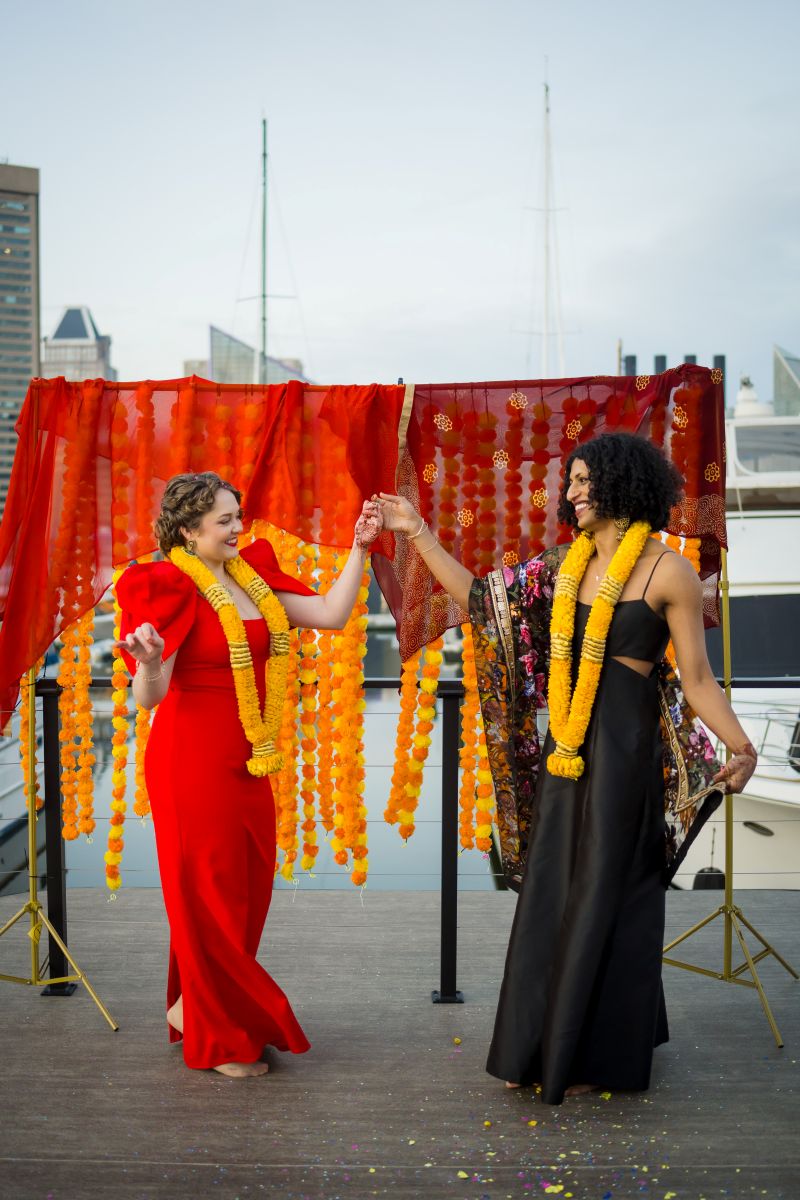 two women dancing with other another and holding hands one woman is wearing a red dress and yellow lei the other woman is wearing a black dress and yellow lei 