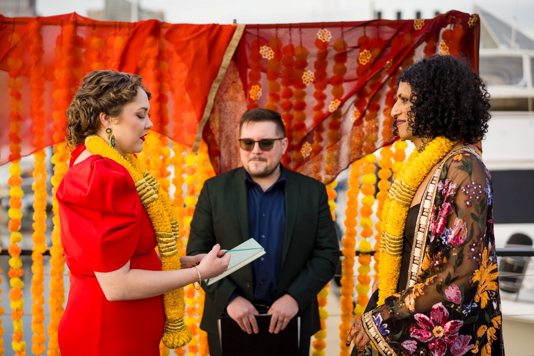 a couple at their wedding ceremony with their offiicant one woman is wearing a red dress and yellow lei and is reading vows to her parter who is wearing a yellow lei and colorful kimono 