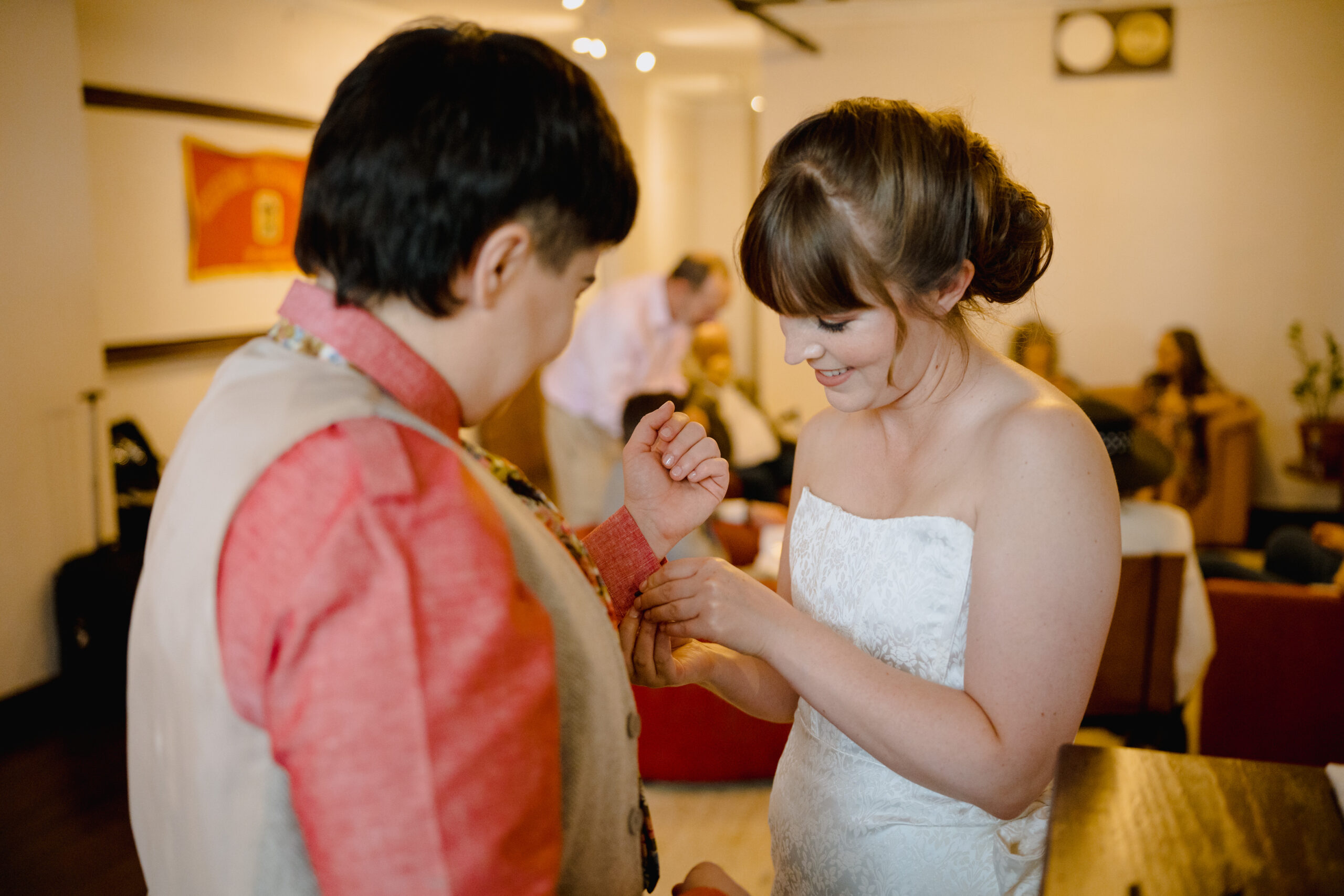 Woman in white dress helps partner in vest and red button-down shirt with the buttons on their sleeve cuffs while getting ready together before their wedding.