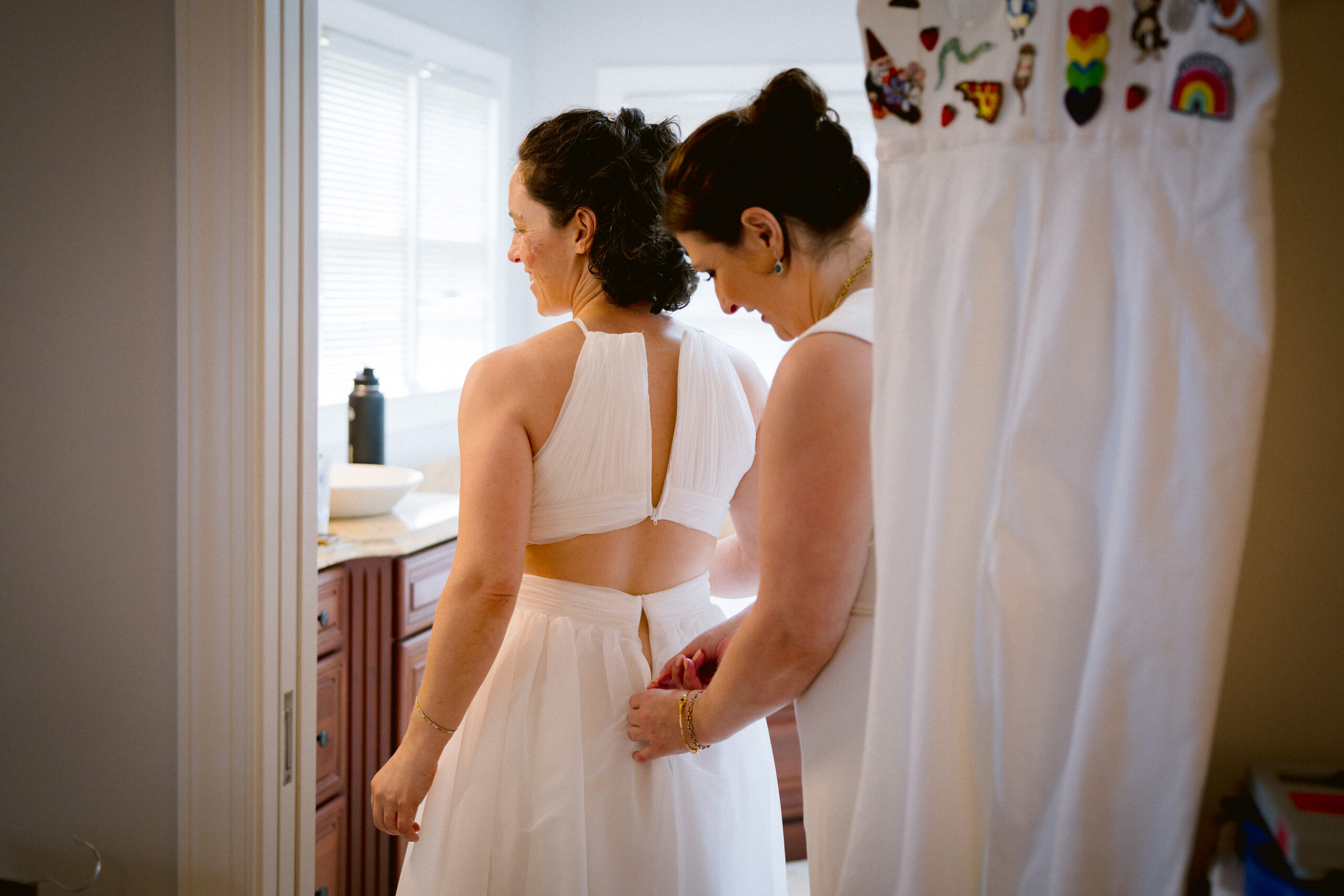 One bride in a white dress zips the other into her white dress while they get ready in their home before their Virginia wedding.