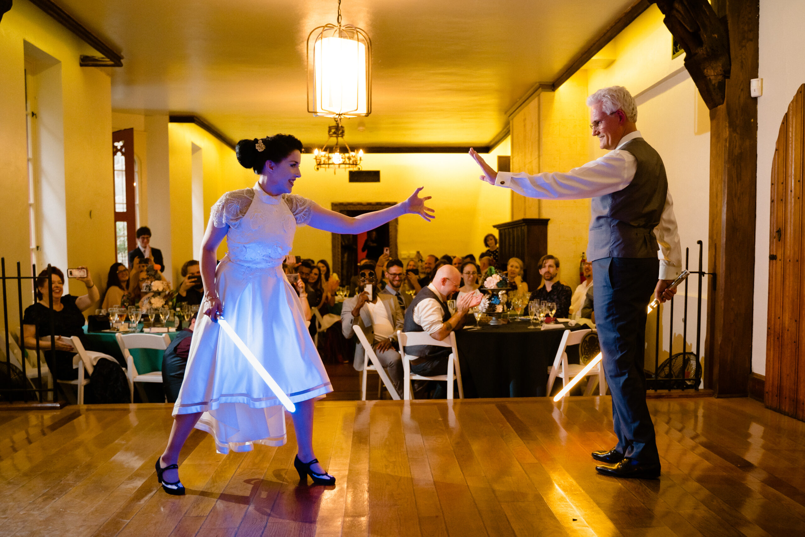 Bride in white dress holds light saber in one hand and gestures at man standing across from her with other hand. Man in suit holds light saber in one hand and the other hand is held out towards the bride. Audience stands and watches this alternative to a father-daughter wedding dance.