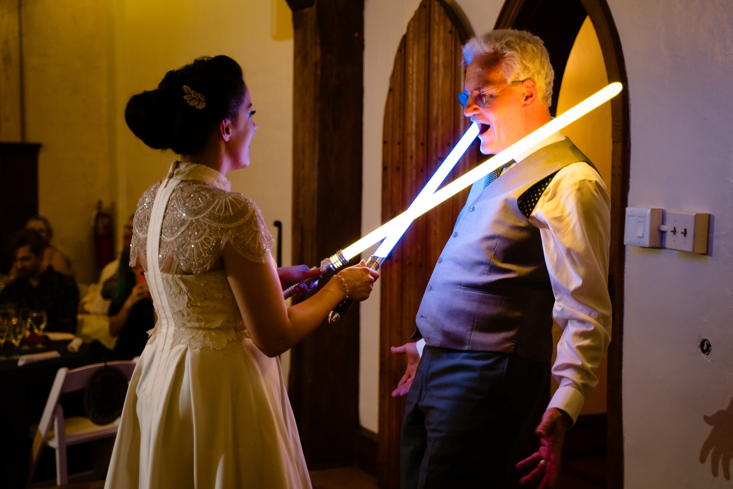 Bride holds two lightsabers crossed in front of her and pointed at her father's neck. Dad gestures with arms out and look of shock on his face.