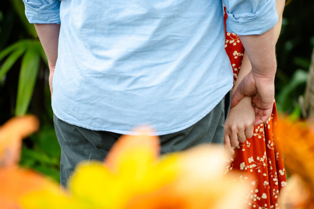Close-up of a couple holding hands during their Rawlings Conservatory engagement photo session in Baltimore, MD, with warm orange flowers softly blurred in the foreground.