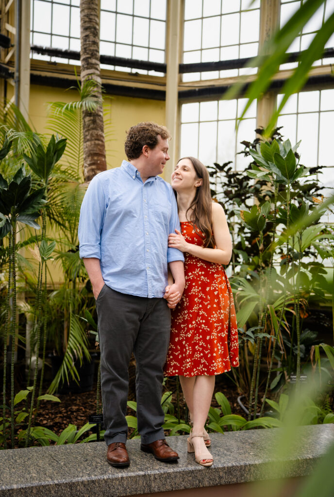 Couple holding hands and smiling inside Rawlings Conservatory in Baltimore, MD, surrounded by tall tropical plants and greenhouse windows during an engagement session.