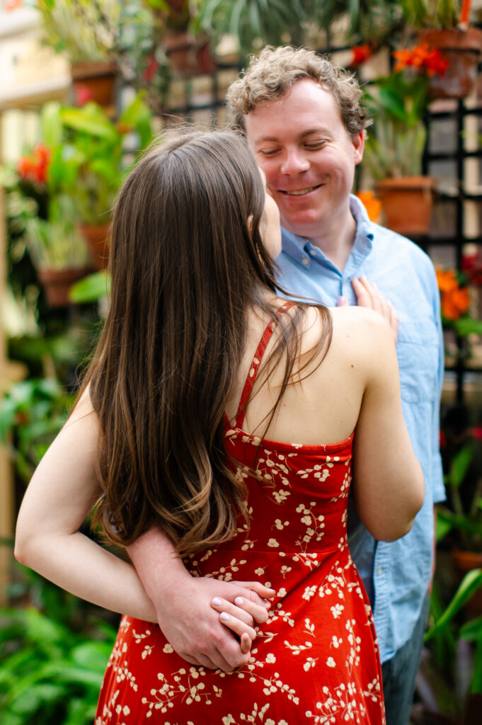 Close-up of a couple embracing during their Rawlings Conservatory engagement photo session in Baltimore, MD, with colorful flowers and greenery softly blurred behind them.