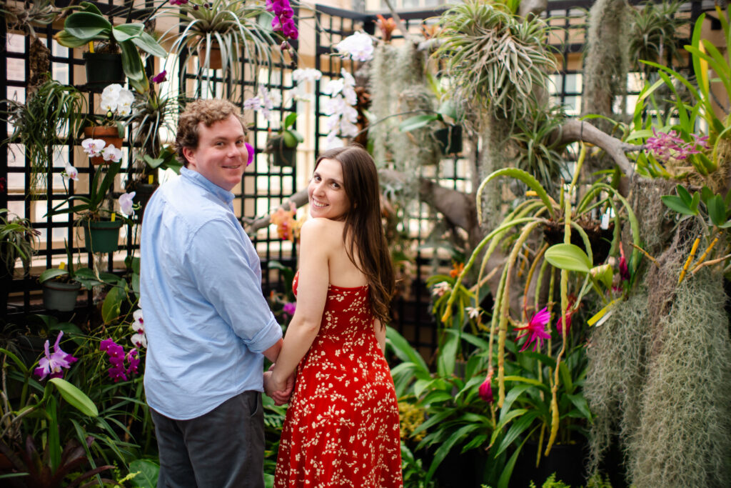 Couple holding hands and looking back at the camera among blooming orchids and lush greenery during their Rawlings Conservatory engagement photo session in Baltimore, MD.