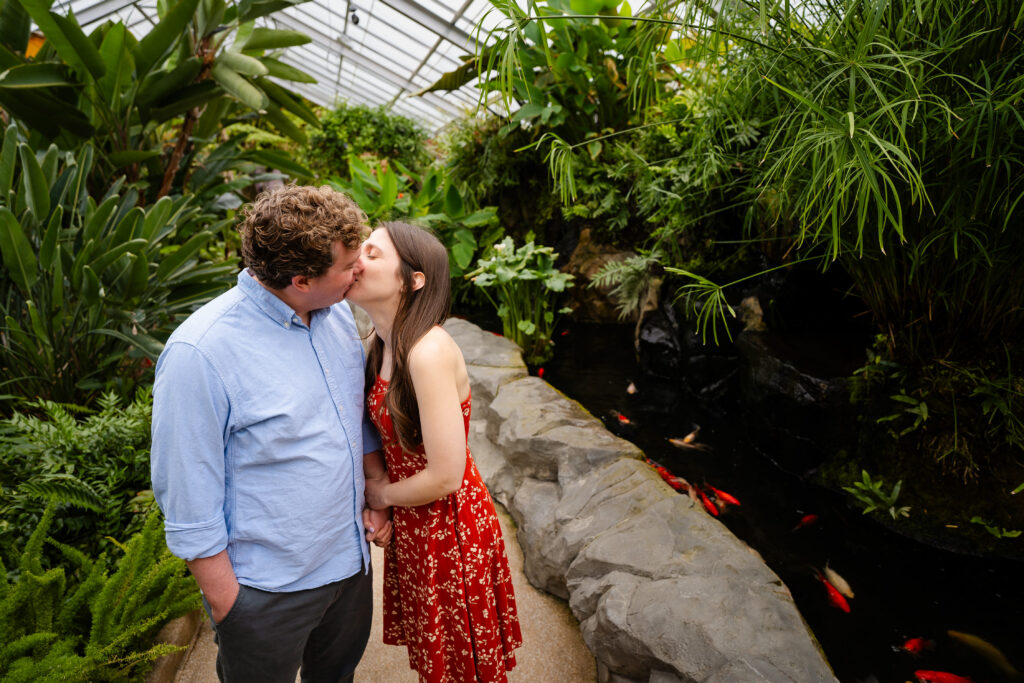 Couple kissing beside the koi pond inside Rawlings Conservatory in Baltimore, MD, surrounded by lush tropical plants during an engagement photo session.