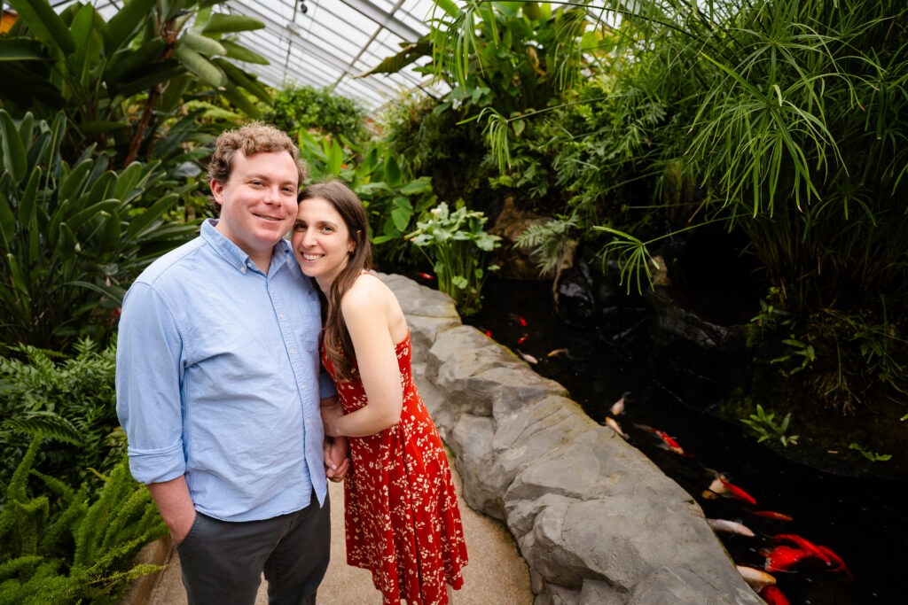 Couple smiling and snuggling by the koi pond at Rawlings Conservatory in Baltimore, MD, with greenhouse greenery and bright orange koi in the water.