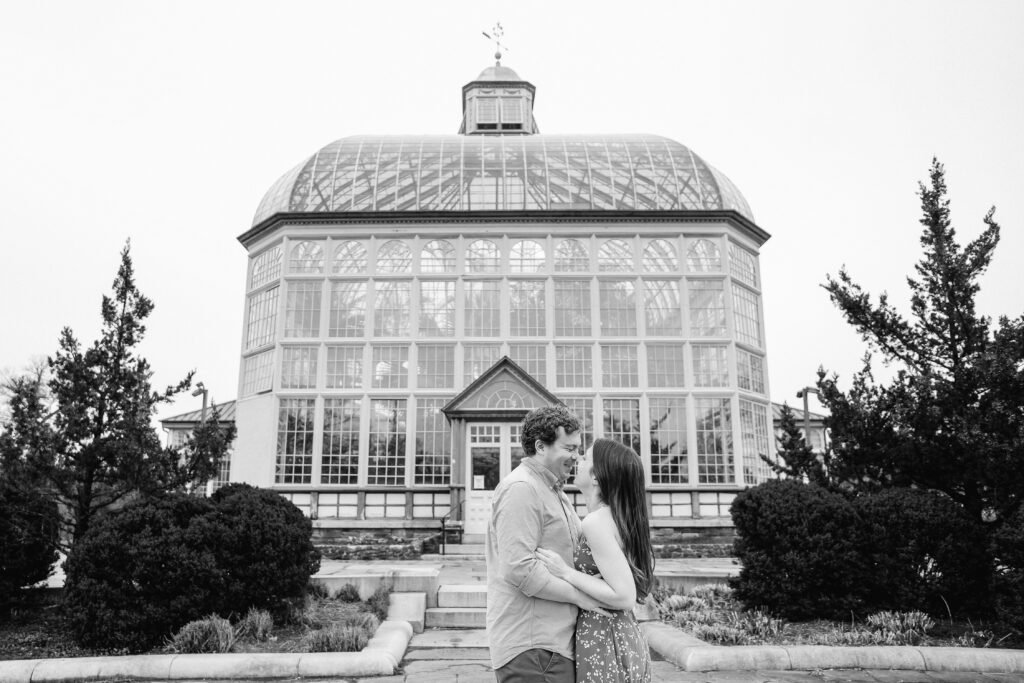 Black-and-white engagement photo of a couple embracing in front of the Rawlings Conservatory greenhouse in Baltimore, MD.